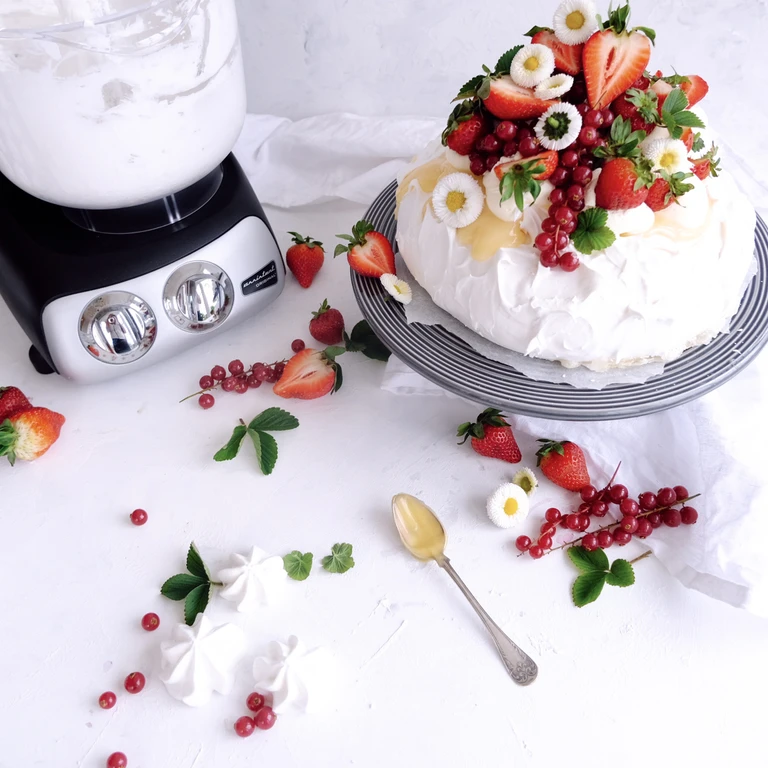 Pavlova cake topped with strawberries and currants beside an Ankarsrum stand mixer with whipped cream bowl.