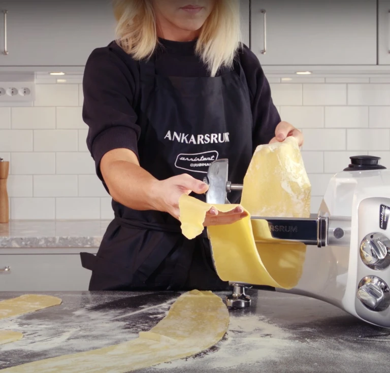 Woman in Ankarsrum apron rolling fresh pasta dough through an Ankarsrum Assistant Original pasta roller attachment.