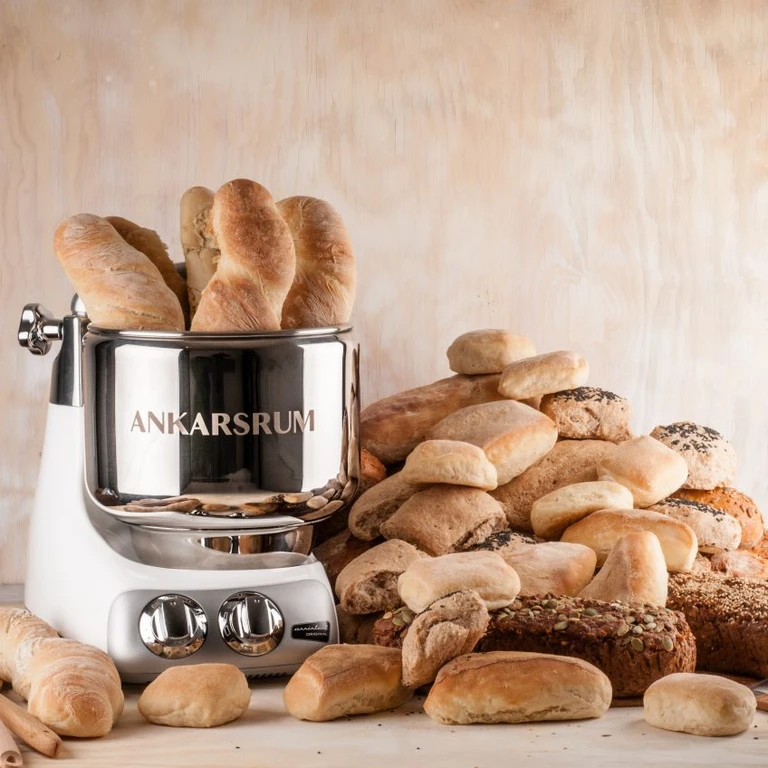 Ankarsrum stand mixer surrounded by freshly baked bread rolls, baguettes, and loaves on a wooden surface.