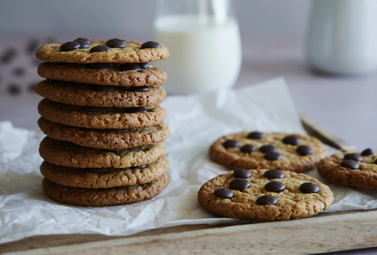 A tall stack of homemade chocolate chip cookies on parchment paper beside a glass of milk, made with an Ankarsrum stand mixer.