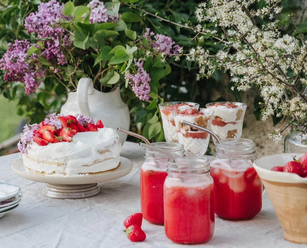 Midsommartårta dekorerad med färska jordgubbar och blommor, serverad på ett vitt bord i en solig trädgård.