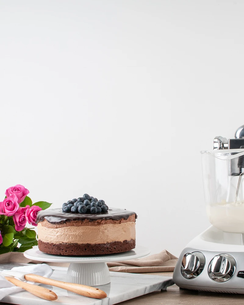 Chocolate mousse cake topped with blueberries on a cake stand beside an Ankarsrum stand mixer and pink roses.