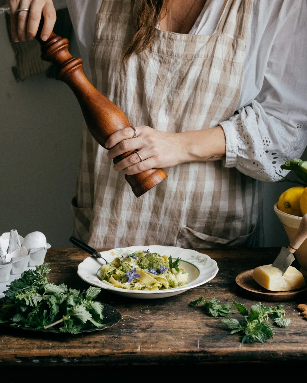 Person in linen apron grinding pepper over fresh nettle pesto pasta with parmesan, lemons, and eggs on rustic wooden table.