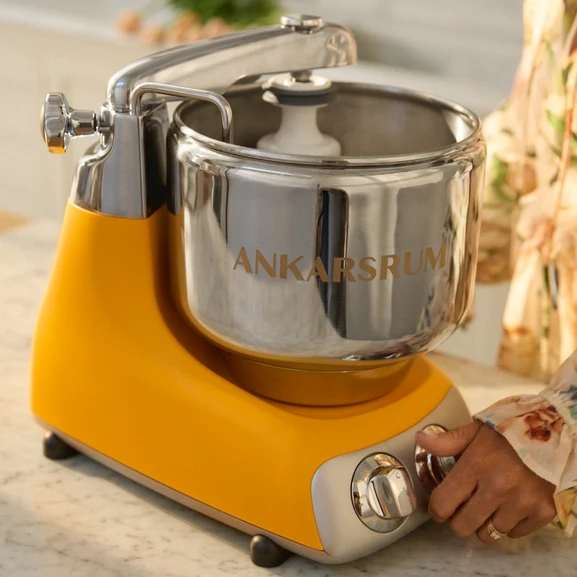 Close-up of the Ankarsrum Assistent Original in Sunbeam Yellow, with a person’s hand resting on the machine.