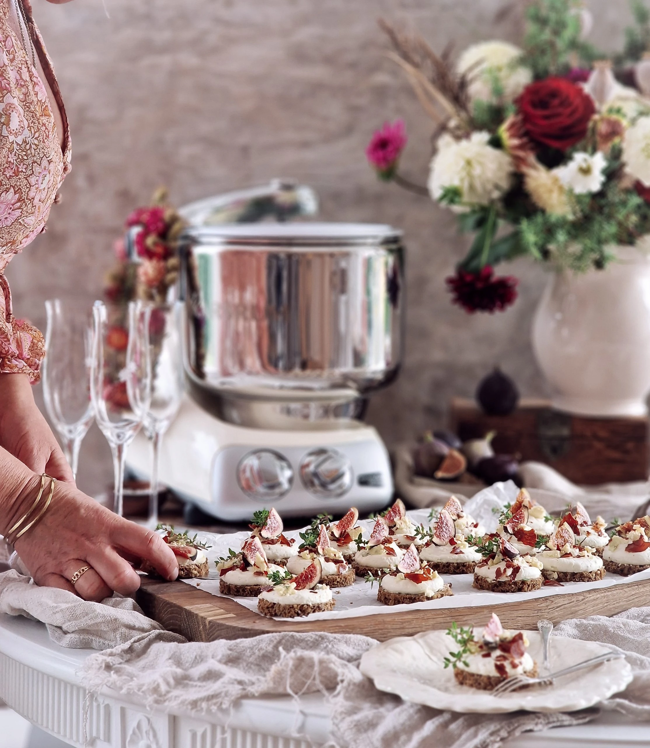 Woman arranging elegant fig and cream cheese canapés on a wooden board beside a silver Ankarsrum stand mixer.