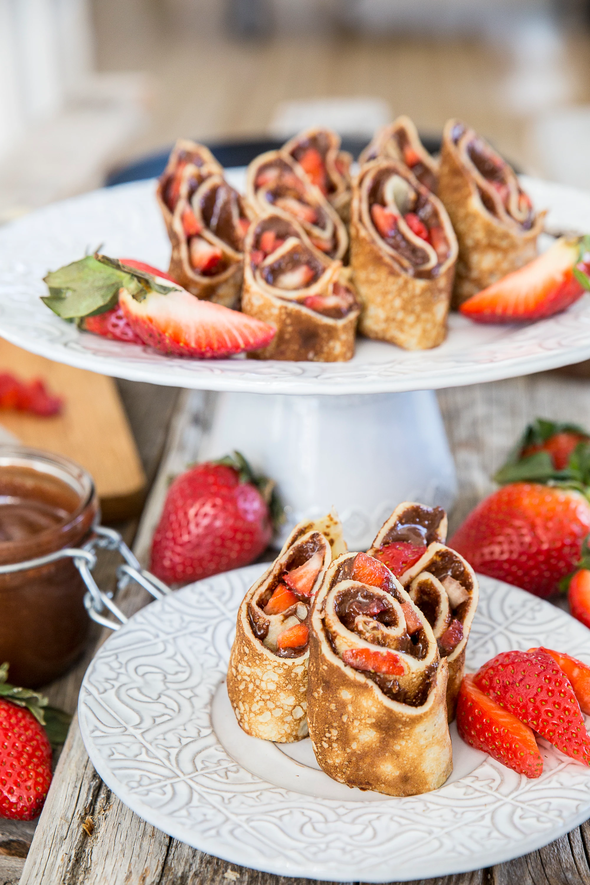 Rolled crepes filled with Nutella and fresh strawberries, displayed on white decorative plates with a jar of chocolate spread nearby.