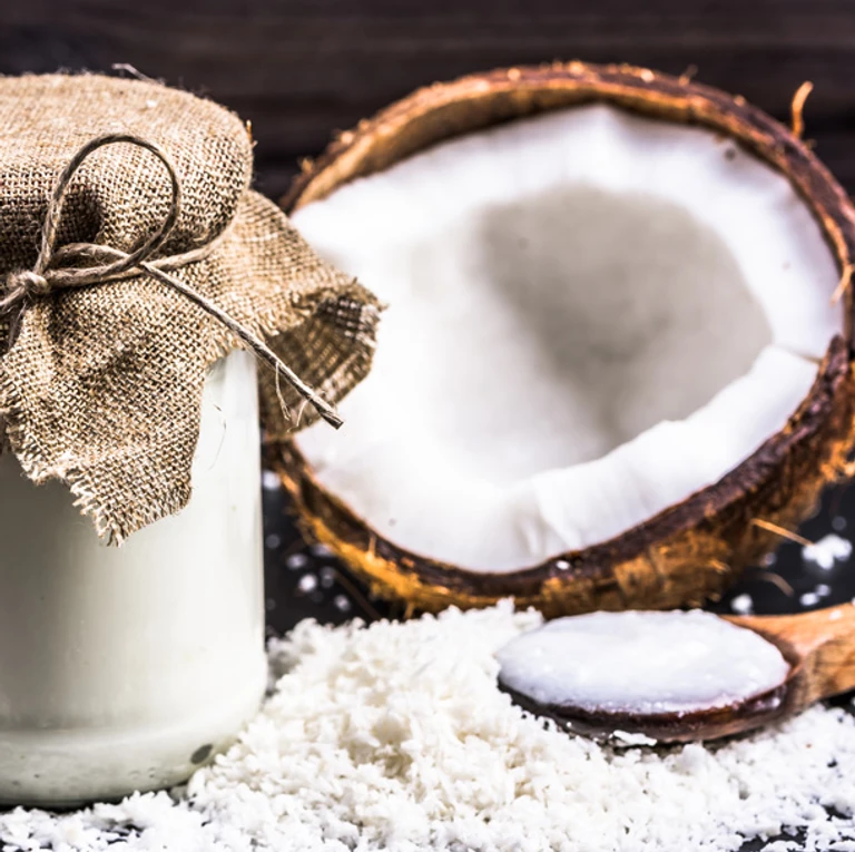Halved coconut with shredded coconut flakes and a rustic jar of coconut milk on a dark wooden surface.