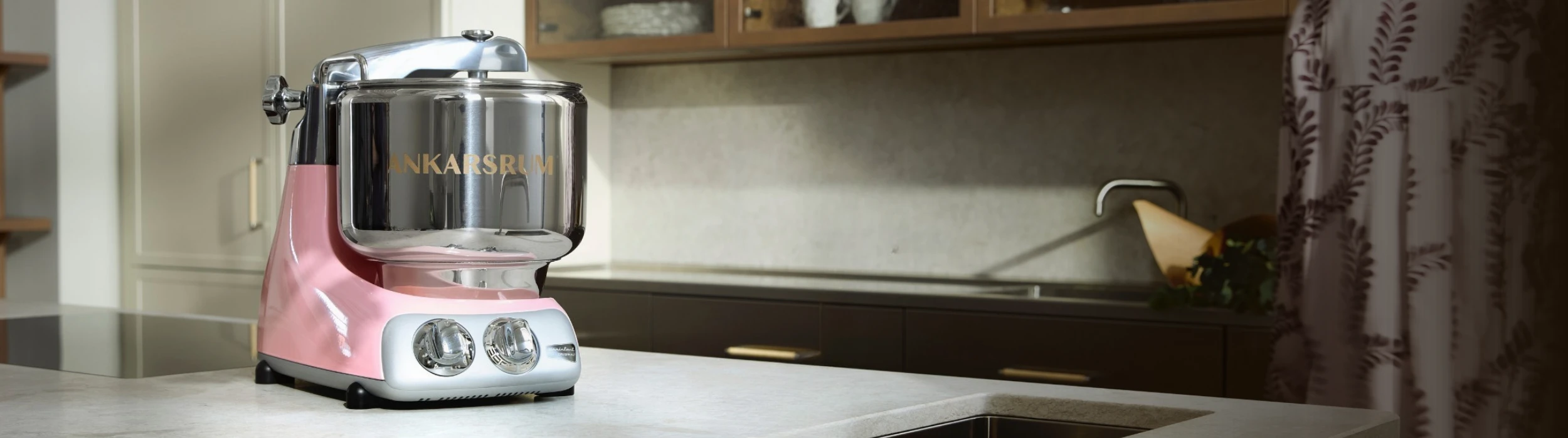 Ankarsrum stand mixer in pink with chrome bowl on a marble kitchen countertop in an elegant modern kitchen.