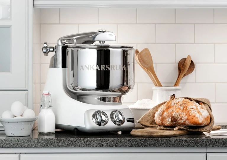 Ankarsrum stand mixer in white and chrome on a kitchen counter with fresh bread, eggs, milk, flour, and wooden utensils.