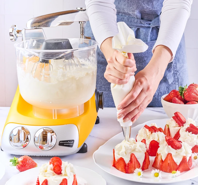 Ankarsrum Assistent Original sunbeam yellow stand mixer whipping cream with balloon whisk attachment, used to decorate a strawberry cake with a piping bag.
