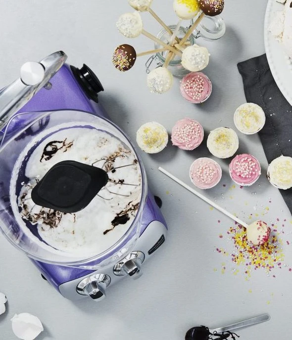 Ankarsrum purple stand mixer bowl with white batter beside colorful cake pops and decorated cupcakes on a gray surface.