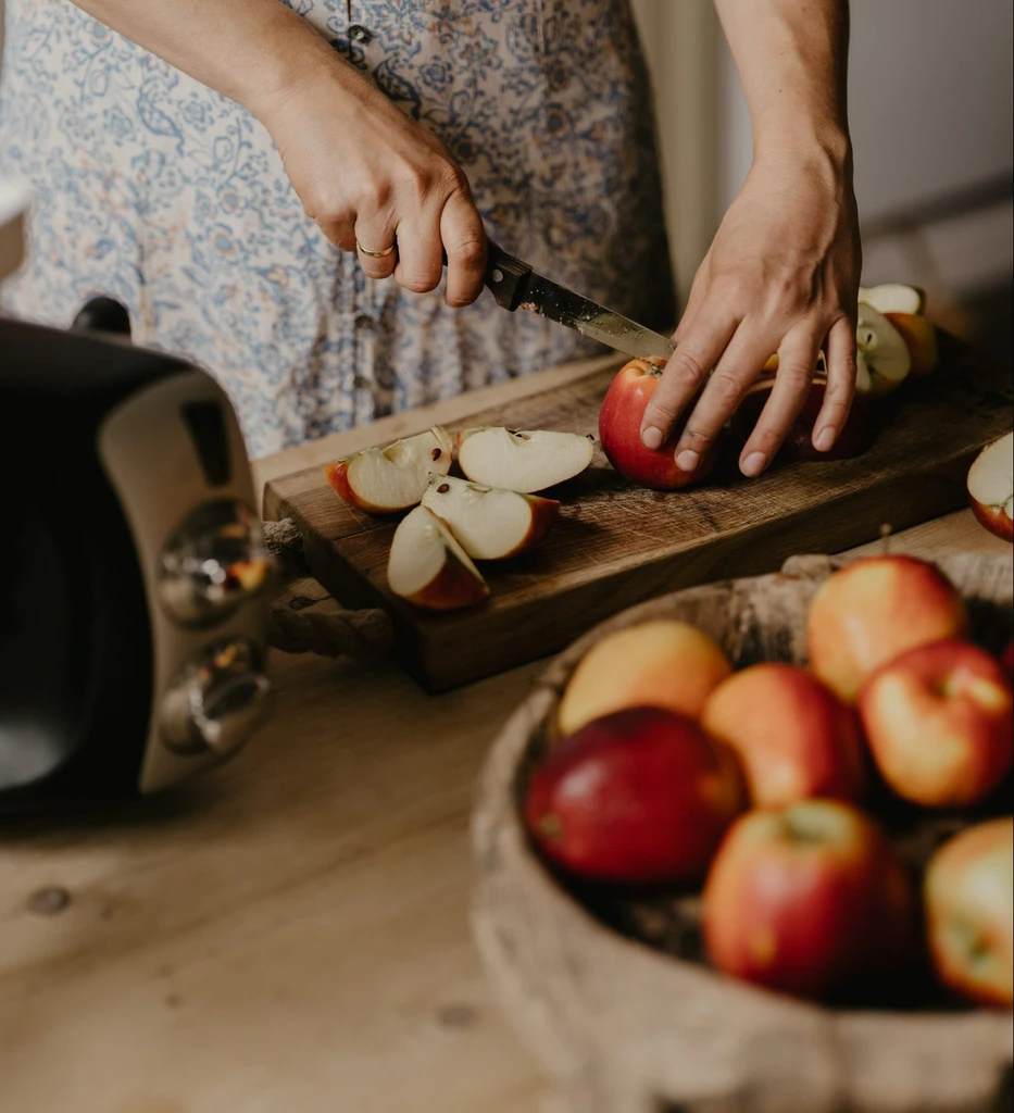 Person slicing fresh red apples on a wooden cutting board beside an Ankarsrum kitchen mixer.