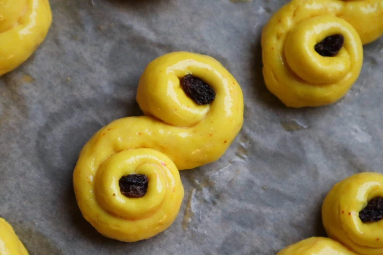 Unbaked saffron buns (lussekatter) with raisins on parchment paper, shaped in traditional S-form, ready for the oven.
