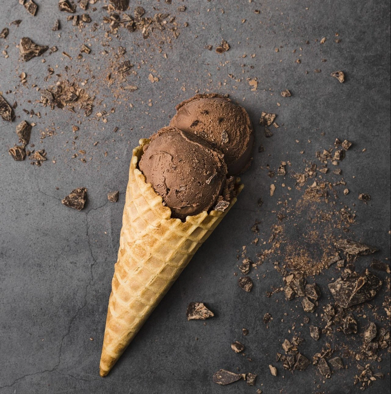 Two scoops of rich chocolate ice cream in a waffle cone, surrounded by chocolate shavings on a dark surface.