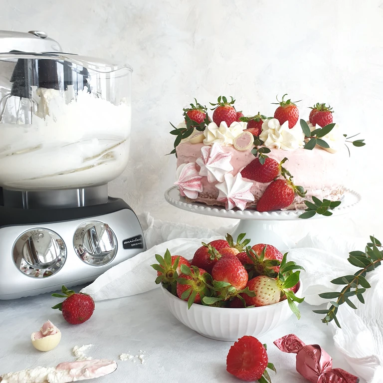Ankarsrum Original stand mixer beside a strawberry cream cake with meringue, fresh strawberries, and whipped cream on a cake stand.