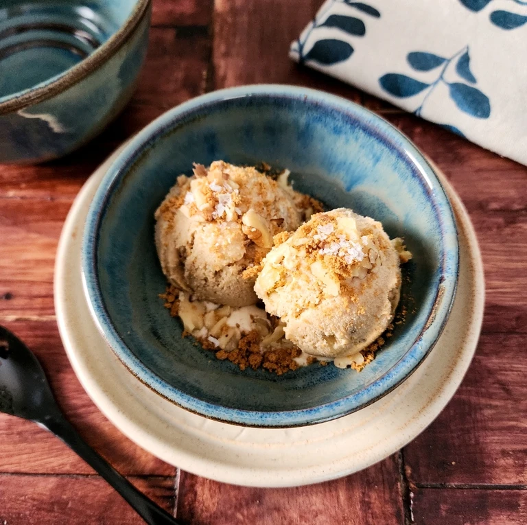 Two scoops of homemade ice cream topped with crumble and sea salt flakes in a blue ceramic bowl.
