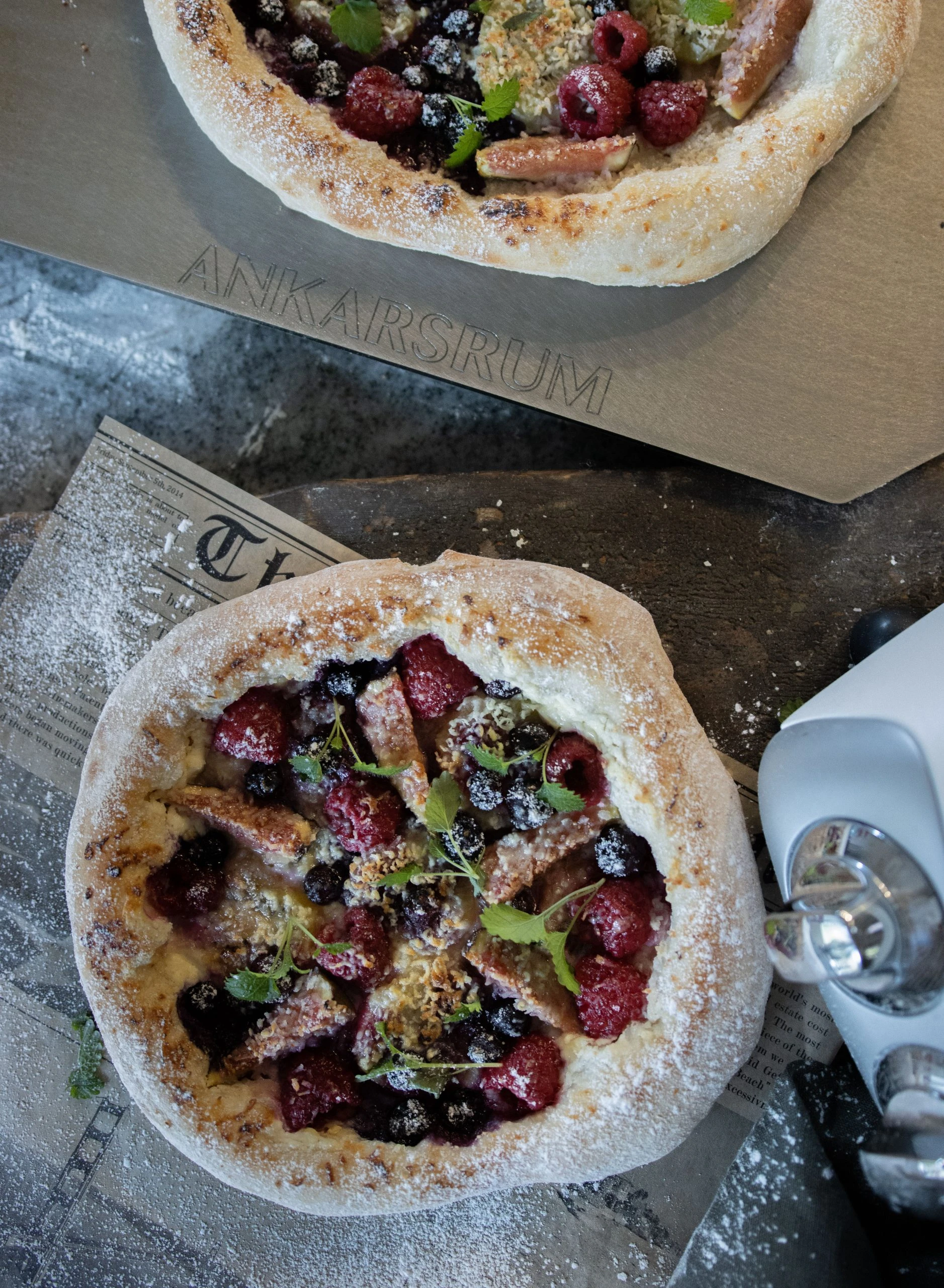 Two rustic berry dessert pizzas dusted with powdered sugar on an Ankarsrum baking stone, topped with raspberries and blueberries.