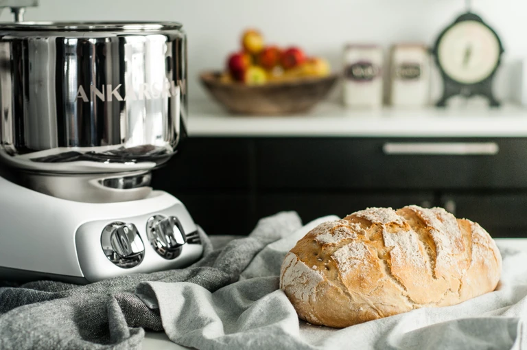 Ankarsrum stand mixer in chrome finish beside a rustic homemade bread loaf on a linen cloth in a kitchen setting.