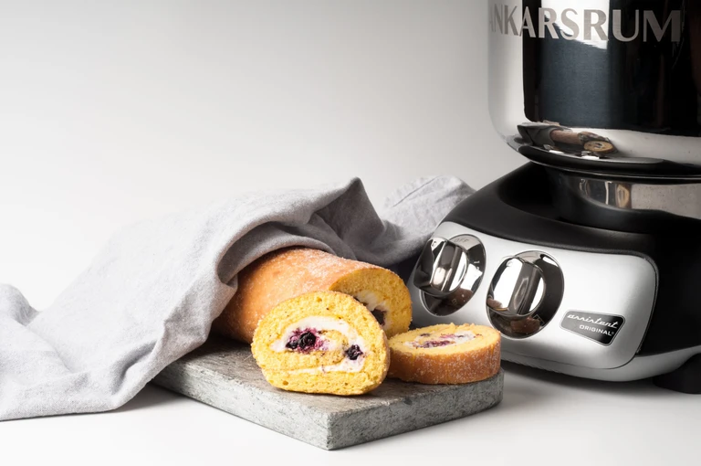 Ankarsrum Assistent Original stand mixer beside a sliced Swiss roll cake with cream and berries filling on a stone board.