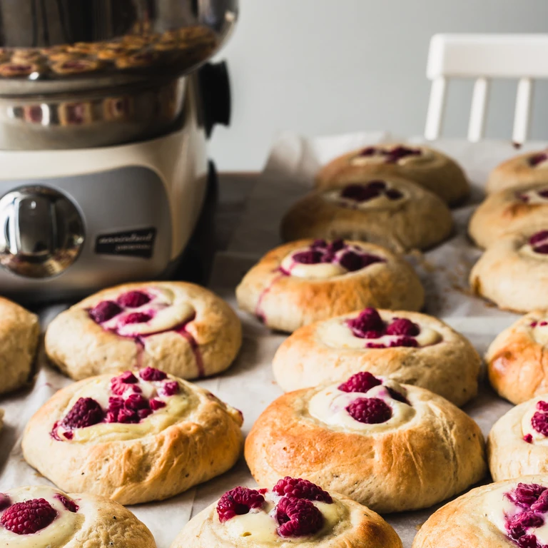 Freshly baked raspberry cream cheese buns on parchment paper beside an Ankarsrum stand mixer.