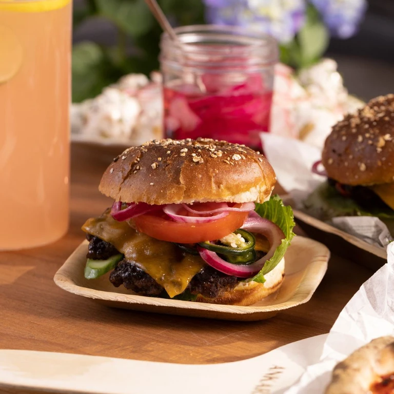 Juicy gourmet burger with melted cheese, jalapeños, pickled red onions, tomato, and lettuce on a seeded bun, served on a wooden plate.