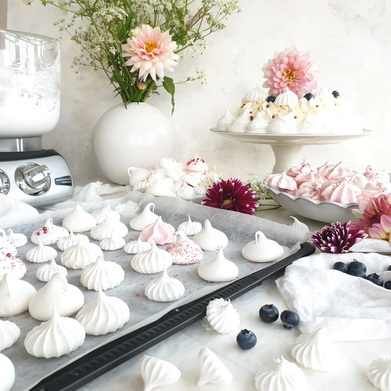 Ankarsrum stand mixer beside trays of homemade white and pink meringues, blueberries, and floral decorations on a marble surface.