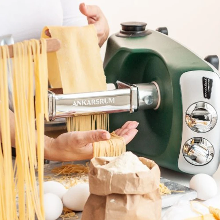 Hands making fresh pasta with Ankarsrum stand mixer pasta attachment, surrounded by flour, eggs, and spaghetti noodles.