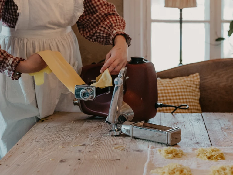 Woman using Ankarsrum stand mixer with pasta roller attachment to roll fresh homemade pasta dough on wooden table.