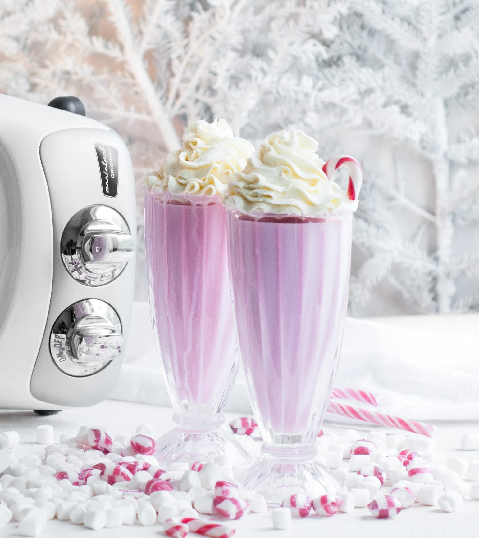Two festive pink peppermint milkshakes topped with whipped cream beside an Ankarsrum Original mixer, surrounded by candy canes and marshmallows.