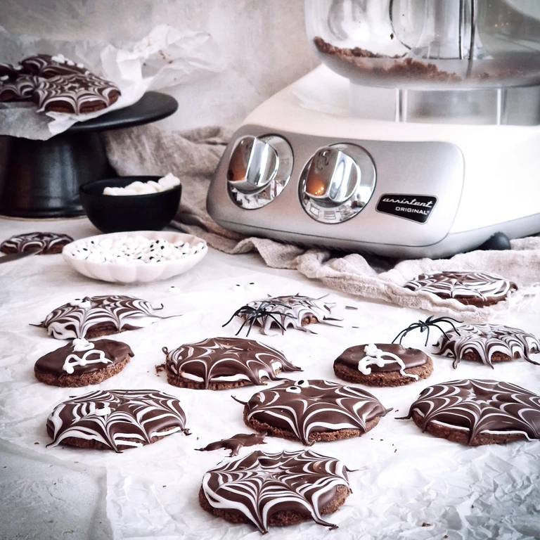 Halloween spider web chocolate cookies displayed beside an Ankarsrum Assistent Original stand mixer with plastic spider decorations.