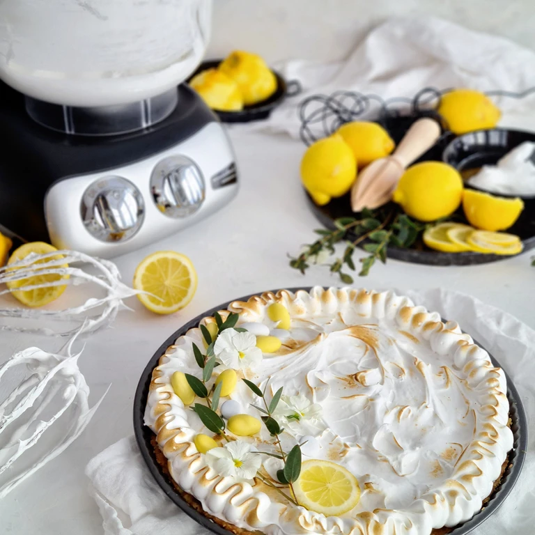 Lemon meringue pie topped with flowers beside an Ankarsrum stand mixer, fresh lemons, and a wooden citrus juicer.