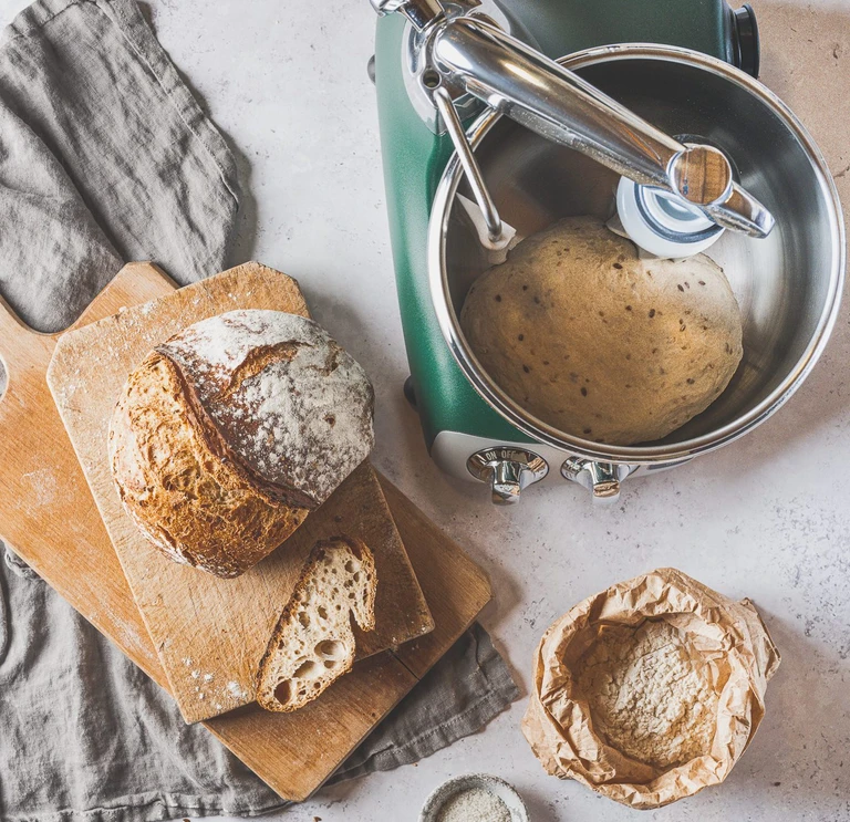 Ankarsrum forest green stand mixer with bread dough in bowl being mixed by the dough roller and dough knife, beside sliced artisan sourdough loaf on wooden cutting board.