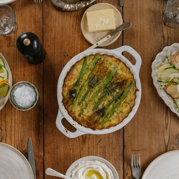 Vårdukning ovanifrån med en sparrisgratäng i mitten, sallader på zucchini, äpple och fänkål runt bordet – lagat med säsongens primörer och serverat i en rustik miljö.