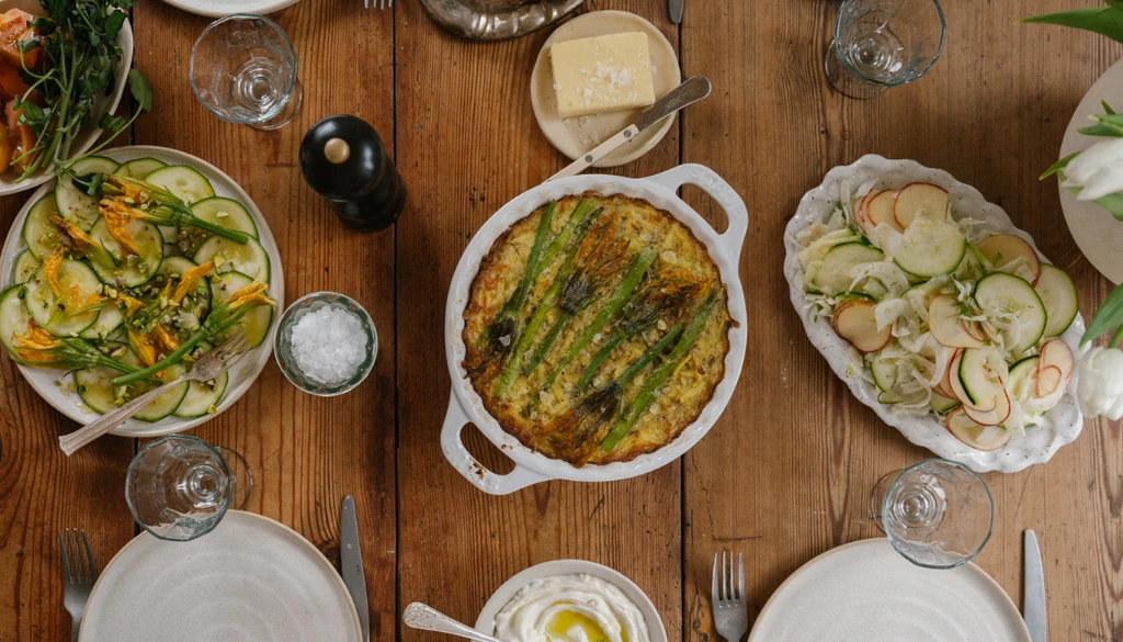 Vårdukning ovanifrån med en sparrisgratäng i mitten, sallader på zucchini, äpple och fänkål runt bordet – lagat med säsongens primörer och serverat i en rustik miljö.
