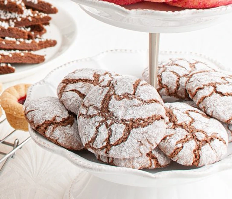 Chocolate crinkle cookies dusted with powdered sugar displayed on a white tiered cake stand with assorted cookies in background.