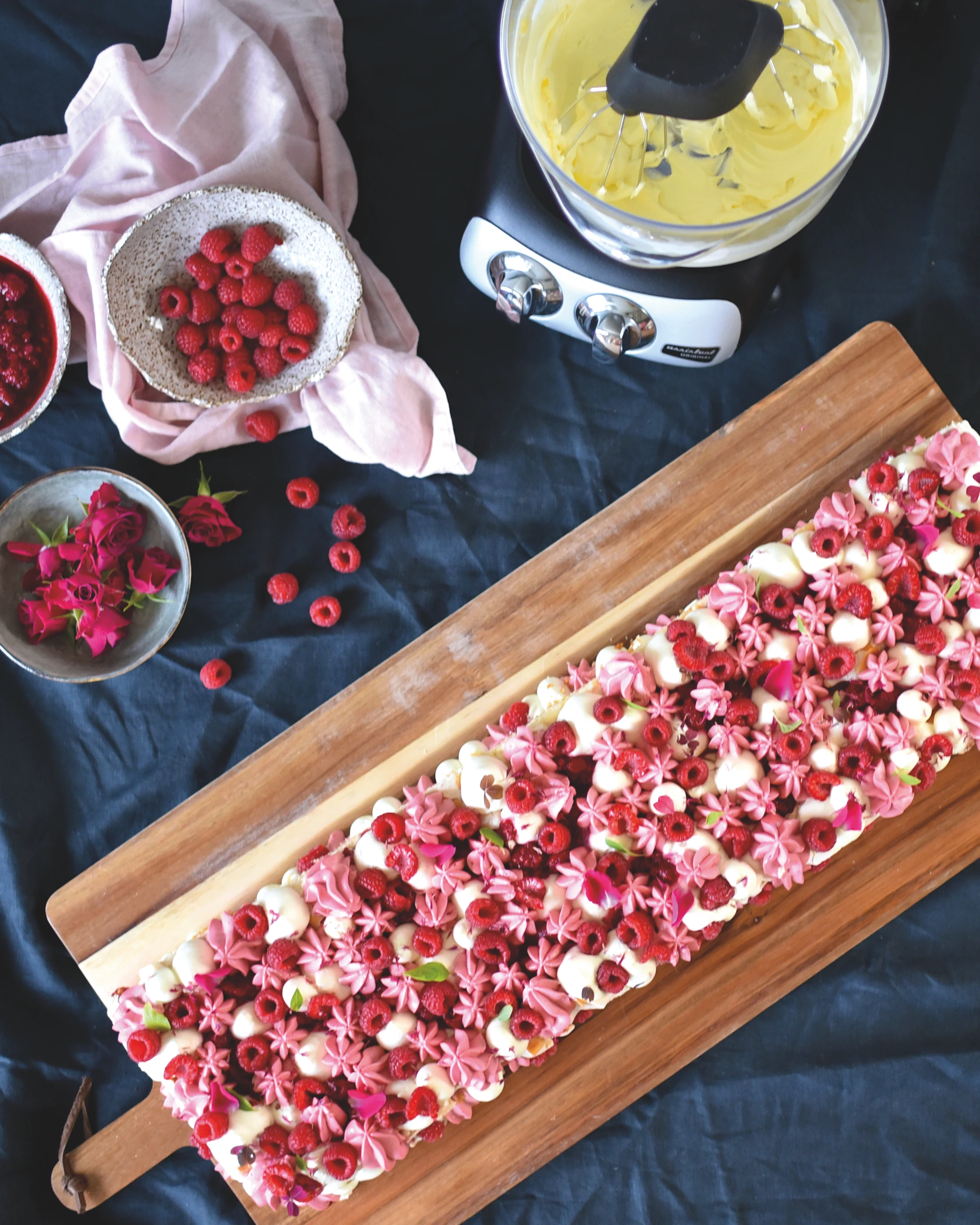 Ankarsrum stand mixer with yellow batter beside a decorated raspberry cake with pink frosting on a wooden board.