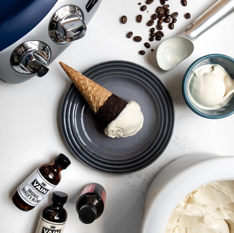 Vanilla ice cream cone on gray plate with VAIN Baker's Blend extract, coffee beans, ice cream scoop, and Ankarsrum mixer.
