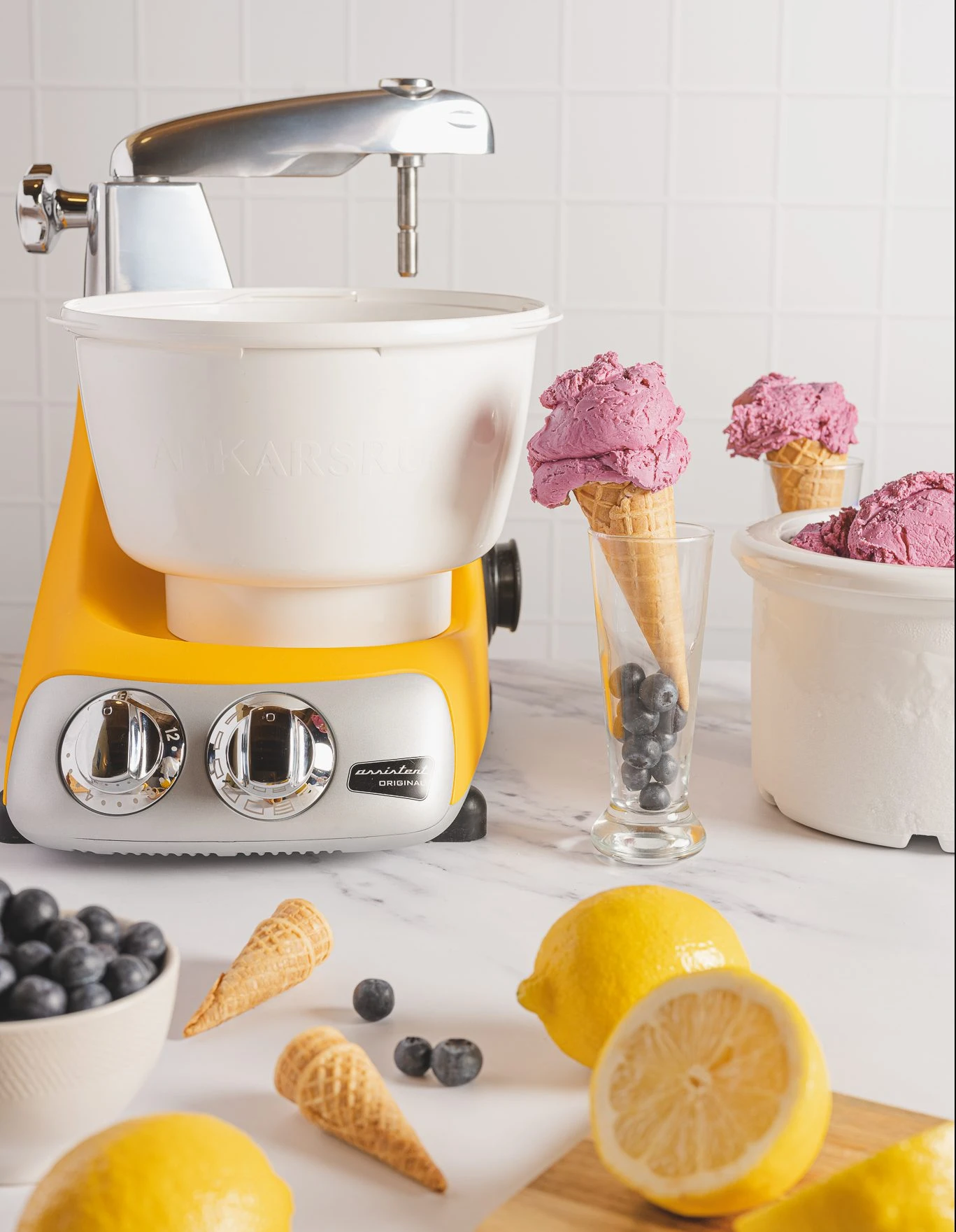 Ankarsrum Original stand mixer in sunbeam yellow with ice cream maker attachment, blueberry ice cream cones, lemons, and fresh blueberries.