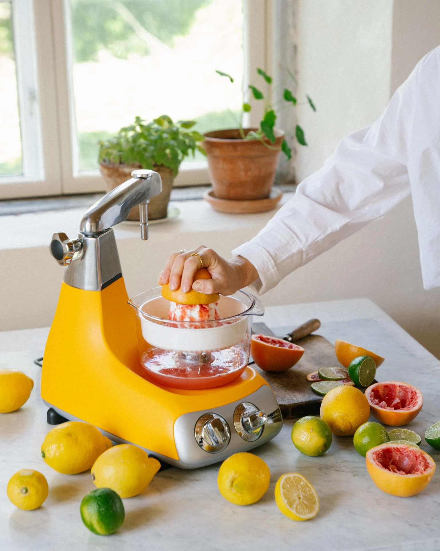 Ankarsrum yellow citrus juicer attachment in use, squeezing fresh grapefruit surrounded by lemons and limes on marble counter.