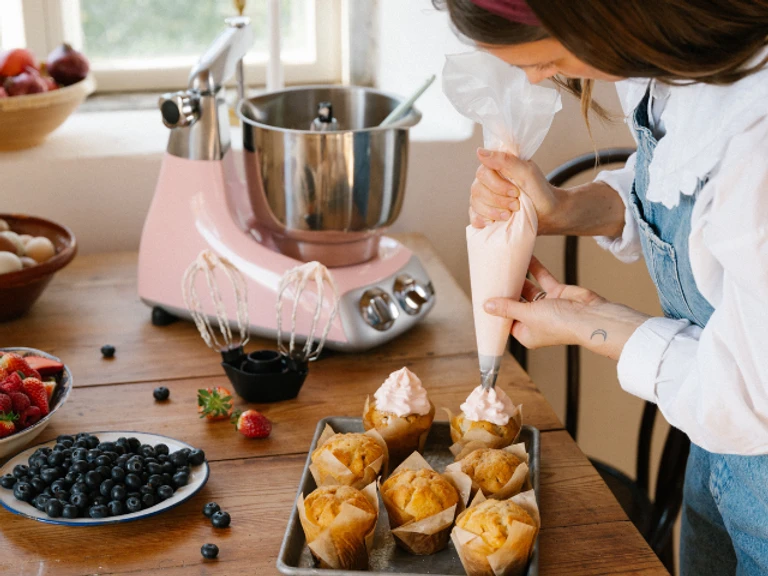 Woman piping pink frosting on muffins beside an Ankarsrum pink stand mixer with balloon whisk attachment on a wooden table.
