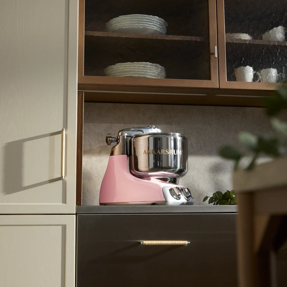 Ankarsrum Assistent Original in Cloudy Pink on a kitchen countertop in a kitchen with dark wood details.