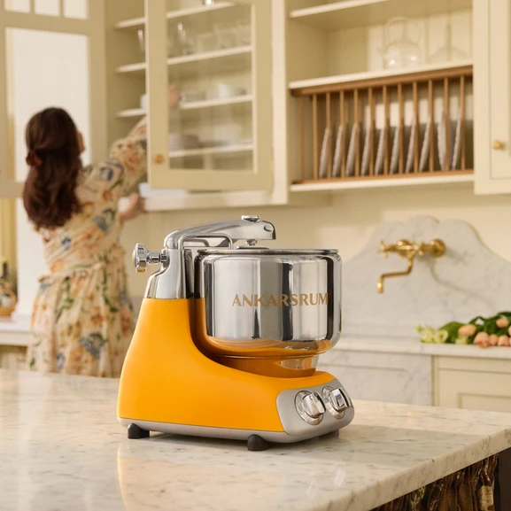 Ankarsrum Assistent Original in Sunbeam Yellow on a marble kitchen island in a bright kitchen, with a person putting items away in a kitchen cabinet in the background.