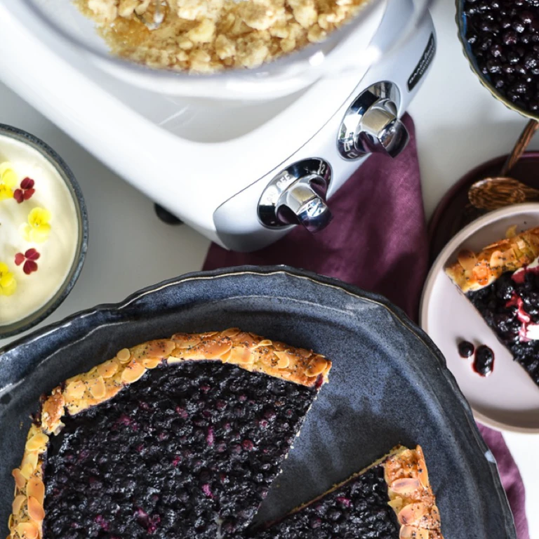 Ankarsrum stand mixer beside a blueberry tart with almond crust, fresh blueberries, cream, and crumble topping on a styled table.