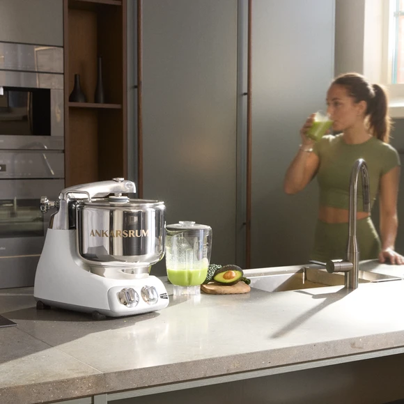 Ankarsrum Assistent Original in Mineral White in a bright kitchen, with a blender filled with green juice beside it and a person drinking a glass of the green juice.