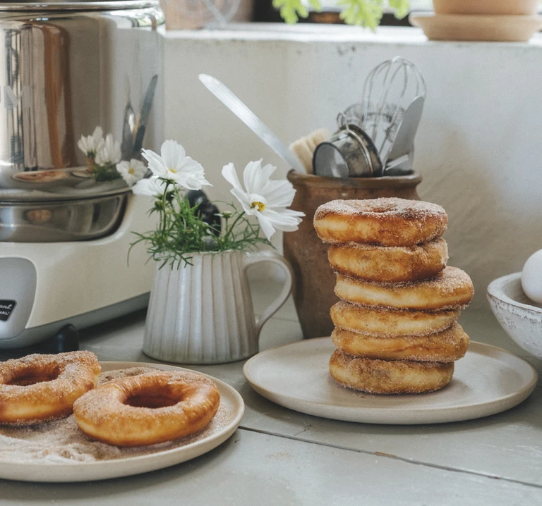 Cinnamon sugar donuts stacked on ceramic plates beside an Ankarsrum stand mixer, white flowers, and kitchen utensils.