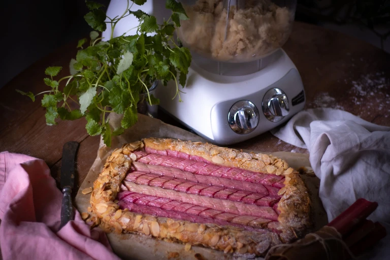 Rustic rhubarb almond galette on parchment paper beside an Ankarsrum stand mixer with dough, fresh herbs in background.