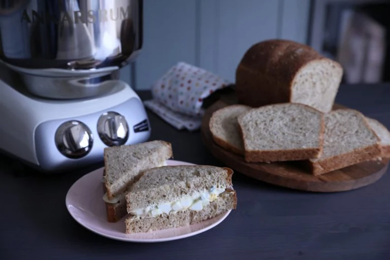 Ankarsrum stand mixer beside sliced homemade whole grain bread loaf and an egg salad sandwich on a pink plate.