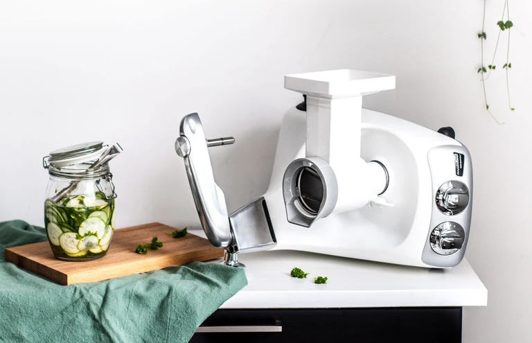 Ankarsrum stand mixer with slicer/shredder attachment beside a jar of sliced cucumbers on a wooden cutting board.