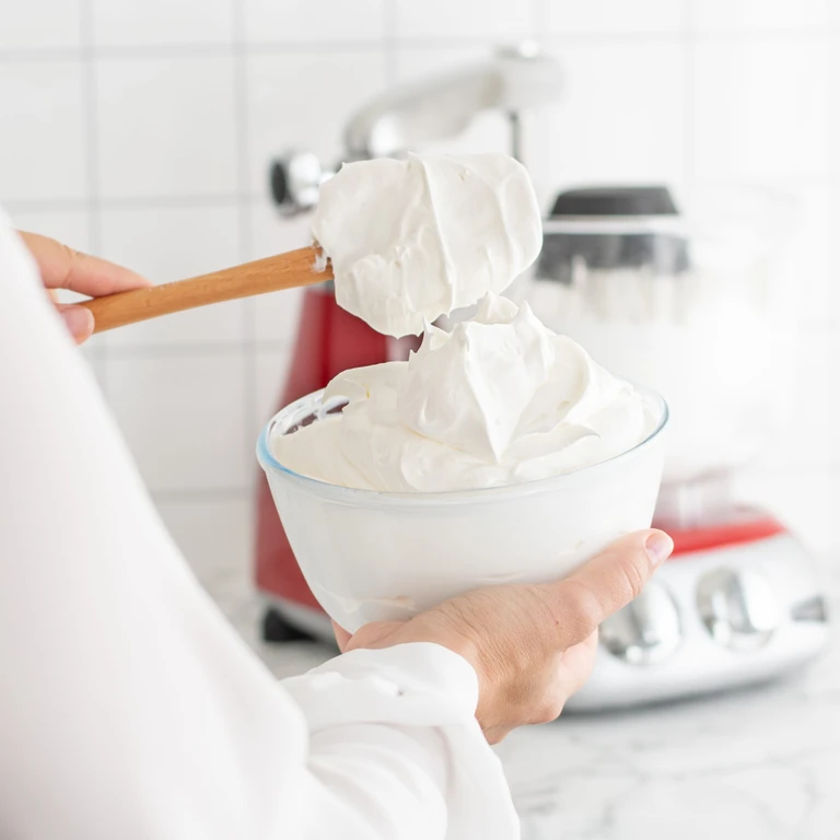 Hands holding a glass bowl of fluffy whipped cream, with a red Ankarsrum stand mixer in the background.