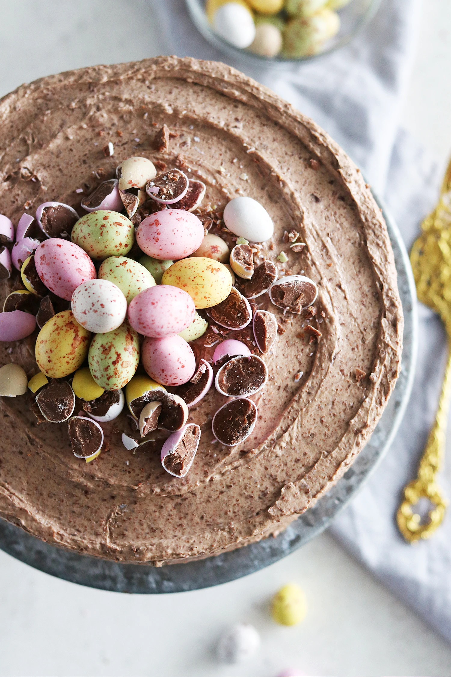 Chocolate Easter cake topped with colorful speckled Mini Eggs and broken candy shell pieces on a gray cake stand.